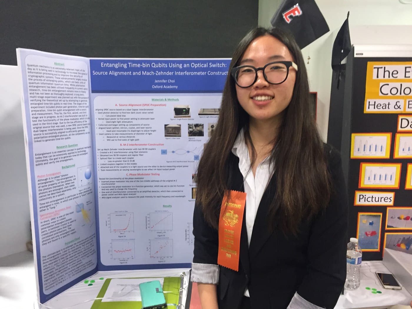 A scholar standing beside her finished display board at the Orange County Science and Engineering Fair, the board carrying an abstract, research question, methods and result charts, with a participant ribbon pinned to her jacket.